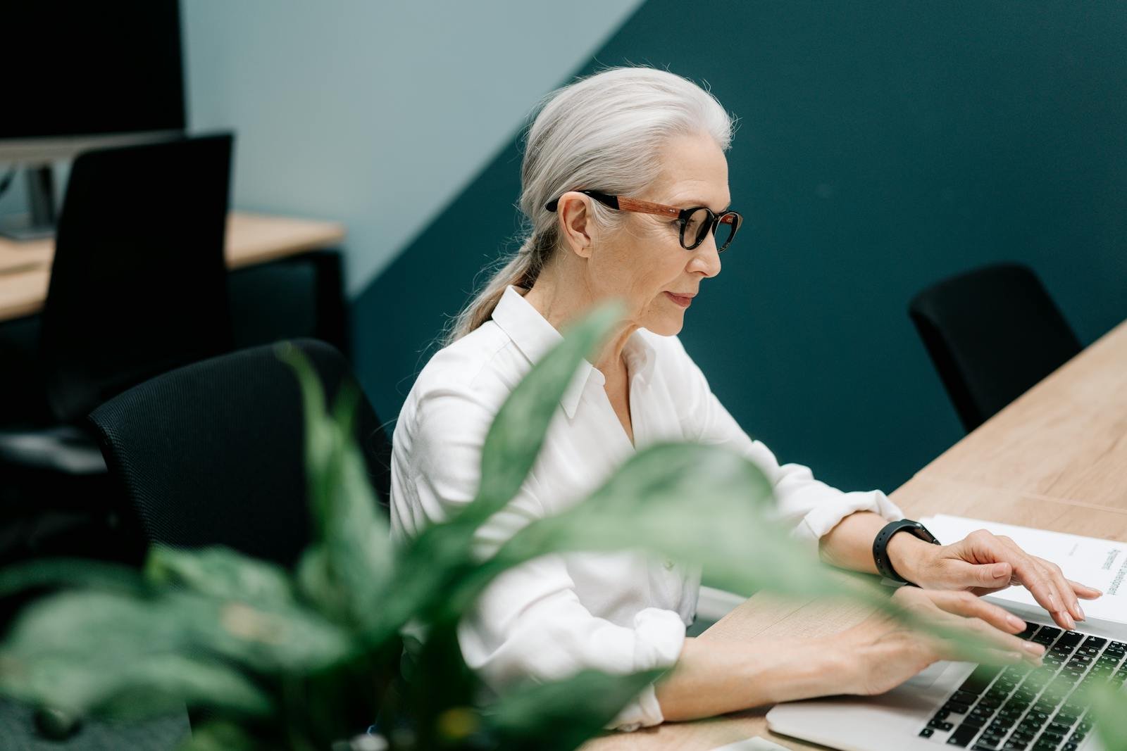 Confident senior woman with glasses working on a laptop in a modern office setting.